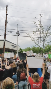 Protestors in Burlington, Vermont raise a "Black Lives Matter" flag on a pole outside the police station. There is a massive crowd and power lines in the foreground.