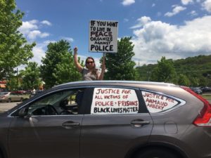 A car is decorated in protest slogans, and a person stand behind the car holding a sign.